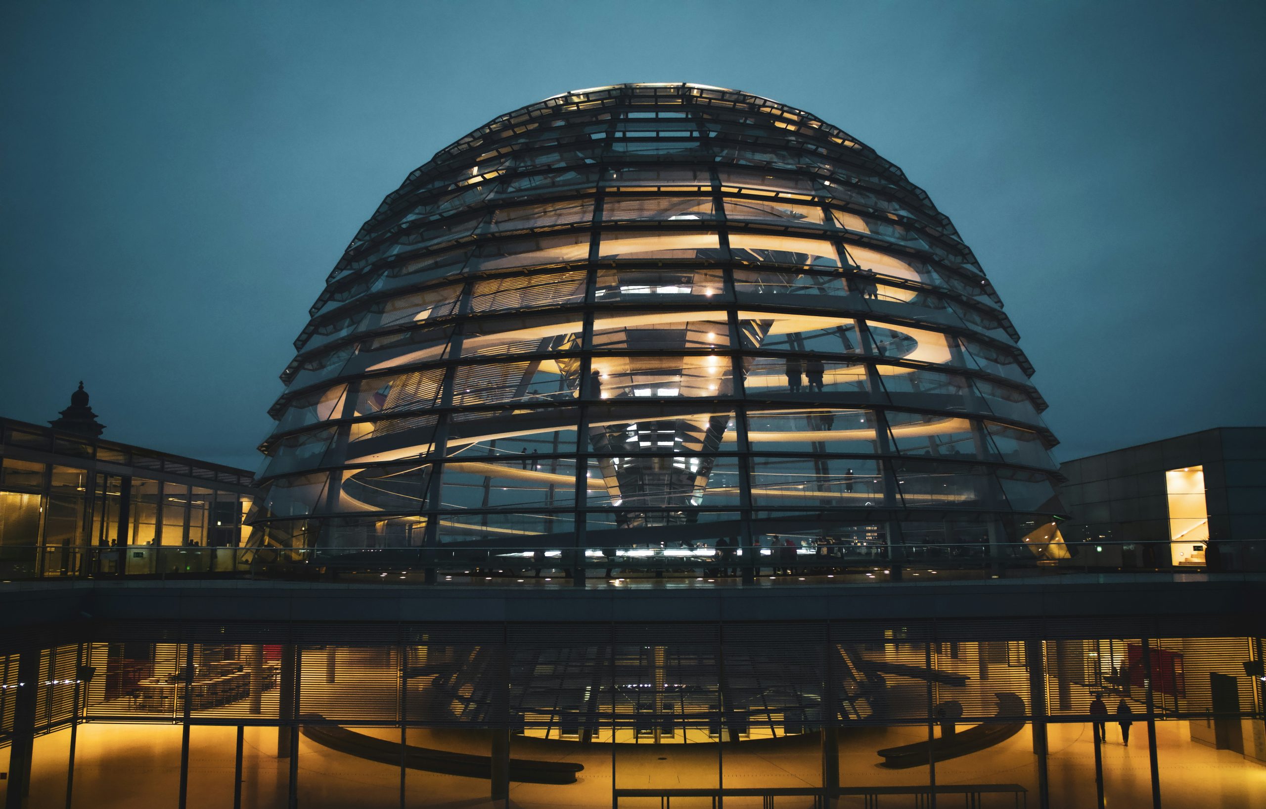 Das Bild zeigt das beleuchtete Reichstagsgebäude in Berlin bei Dämmerung. Die moderne Glaskuppel, ein markantes Merkmal des Gebäudes, ist deutlich sichtbar und beleuchtet, was einen starken Kontrast zur dunklen Umgebung bildet. Der im Reichstagsgebäude tagende Bundestag hat kürzlich einen Gesetzentwurf zur Bürokratieentlastung beschlossen, über den wir in unserem Mandantennewsletter informieren.
